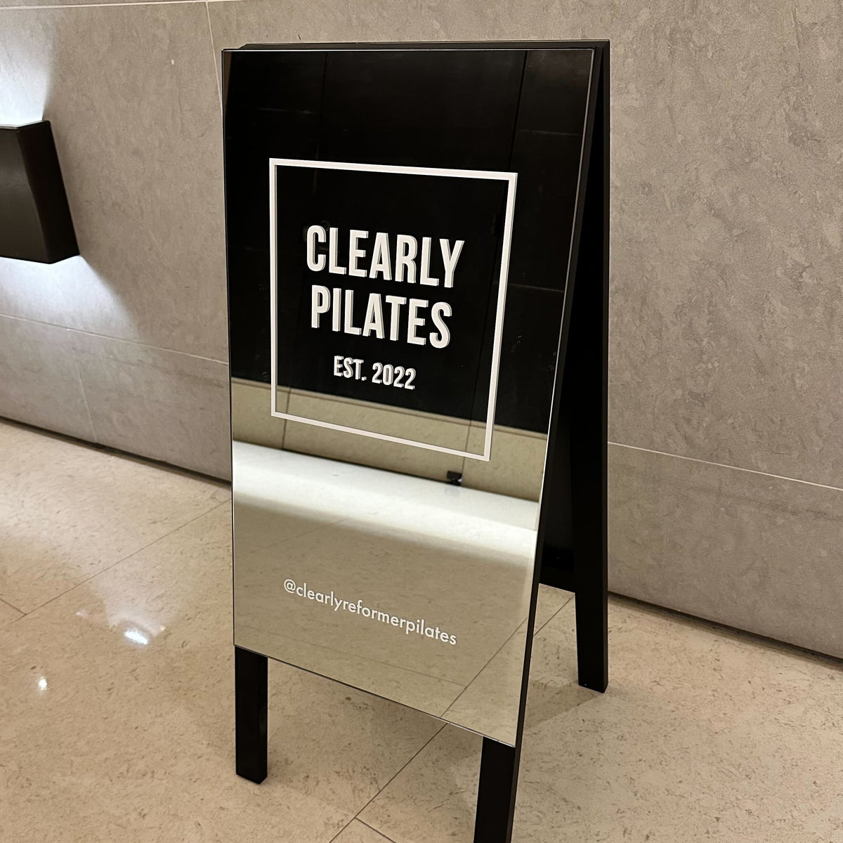Mirrored A-frame sign with a white Clearly Pilates logo and black legs.