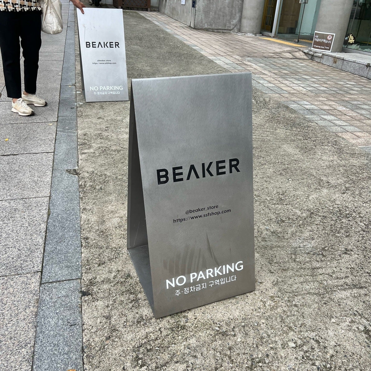 Stainless steel Beaker A-frame sign with a No Parking notice on a sidewalk.