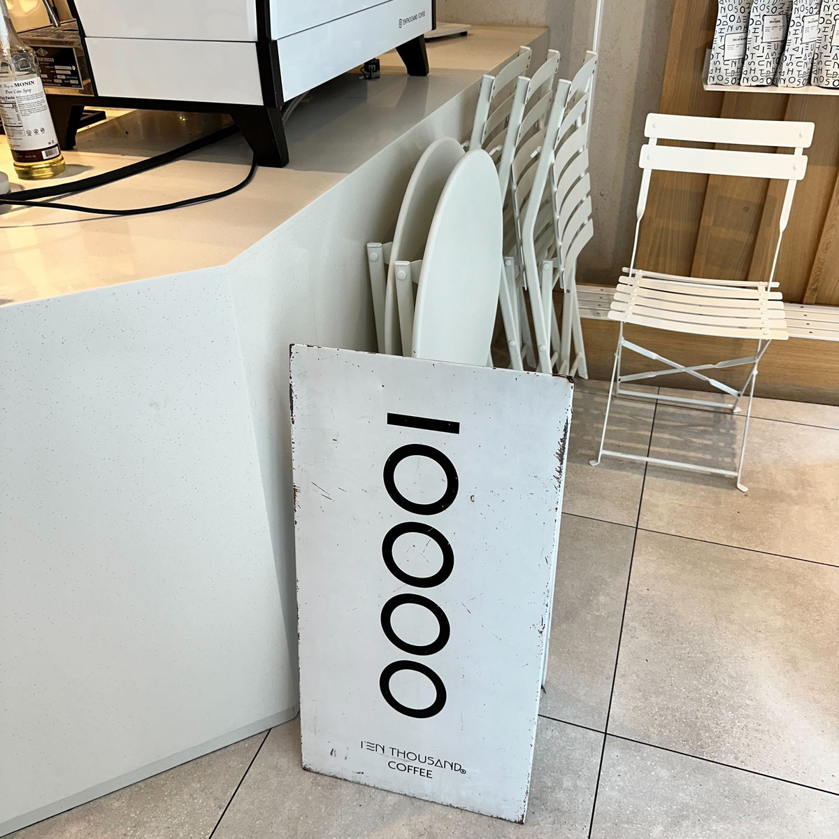 White Ten Thousand Coffee tote bag leaning against a café counter.