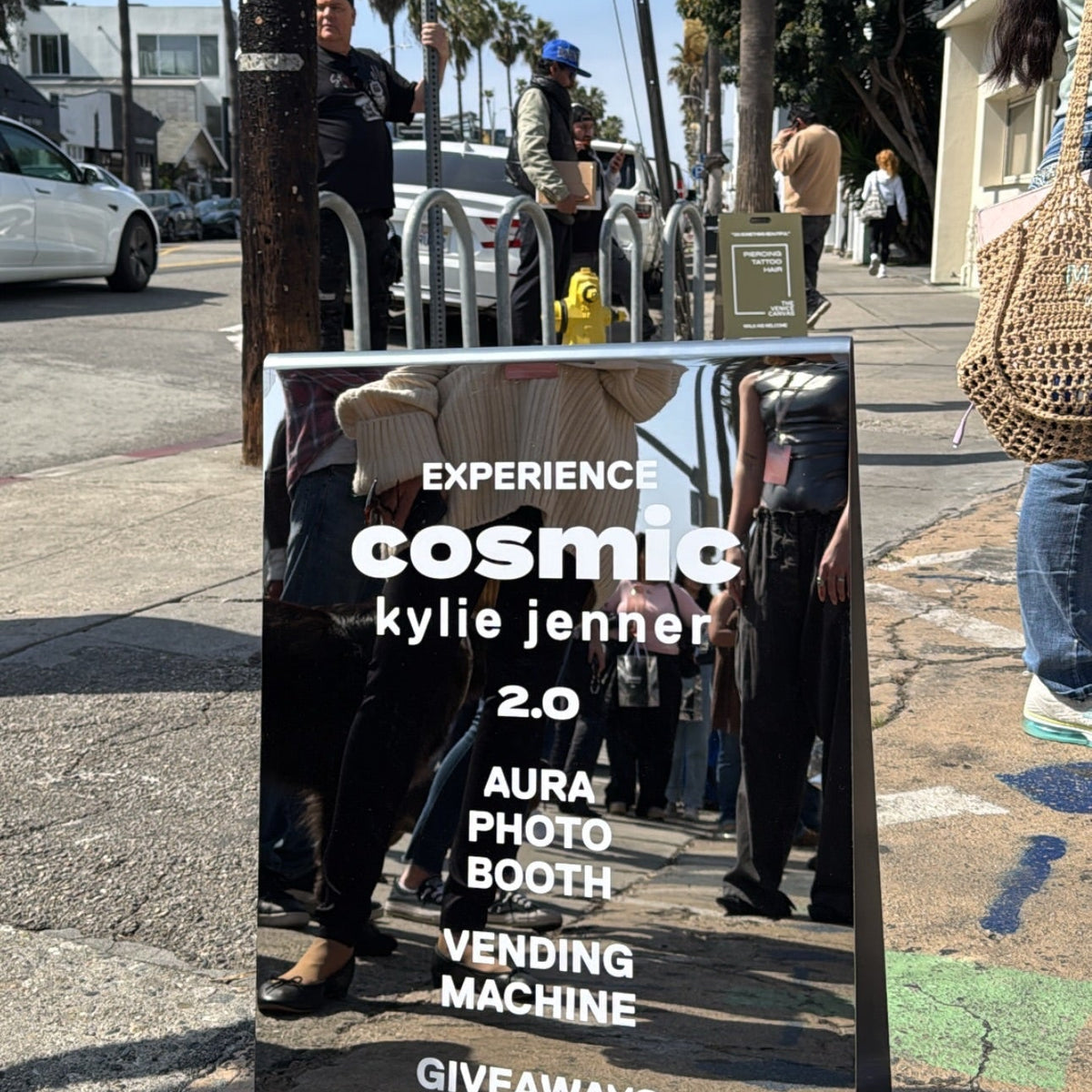 Mirrored A-frame sign listing activities for the Cosmic Kylie Jenner event on a palm-lined sidewalk.