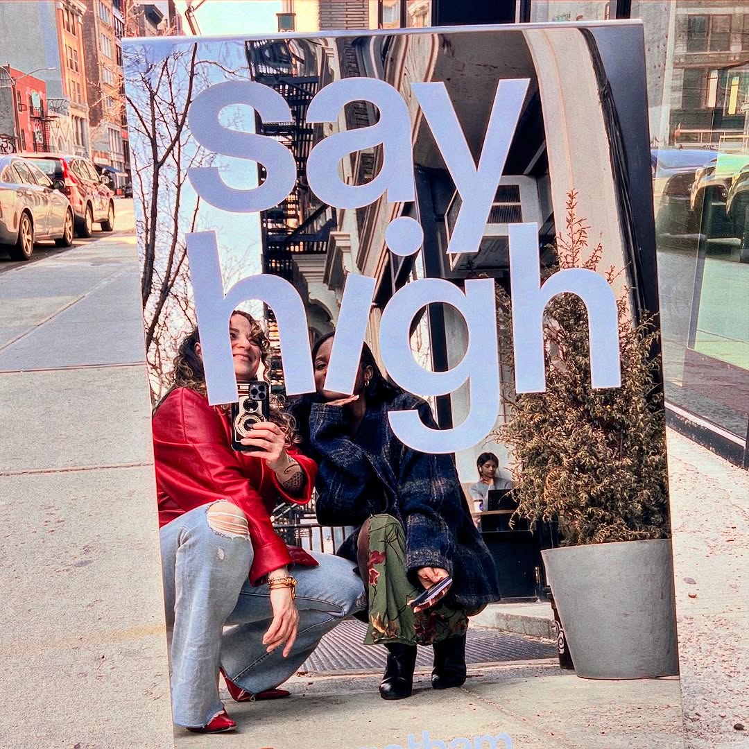 Mirrored Gotham sign with large Say High lettering reflecting people on a sidewalk.