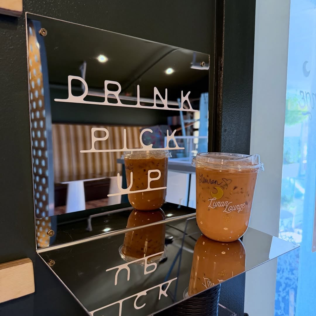 Two iced drinks in clear plastic cups with lids on a mirror shelf, with 'Drink Pick Up' sign in the background.