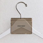 Bronze Smoke acrylic hanger sign clip resting on the rounded crown of a white wooden hanger, with example EVIE PANT print