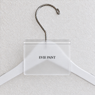 Clear Matte acrylic hanger sign clip resting on the rounded crown of a white wooden hanger, with example EVIE PANT print