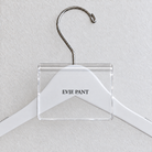Clear acrylic hanger sign clip resting on the rounded crown of a white wooden hanger, with example EVIE PANT print
