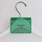 Green acrylic hanger sign clip resting on the rounded crown of a white wooden hanger, with example EVIE PANT print