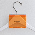 Orange acrylic hanger sign clip resting on the rounded crown of a white wooden hanger, with example EVIE PANT print