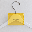 Yellow acrylic hanger sign clip resting on the rounded crown of a white wooden hanger, with example EVIE PANT print