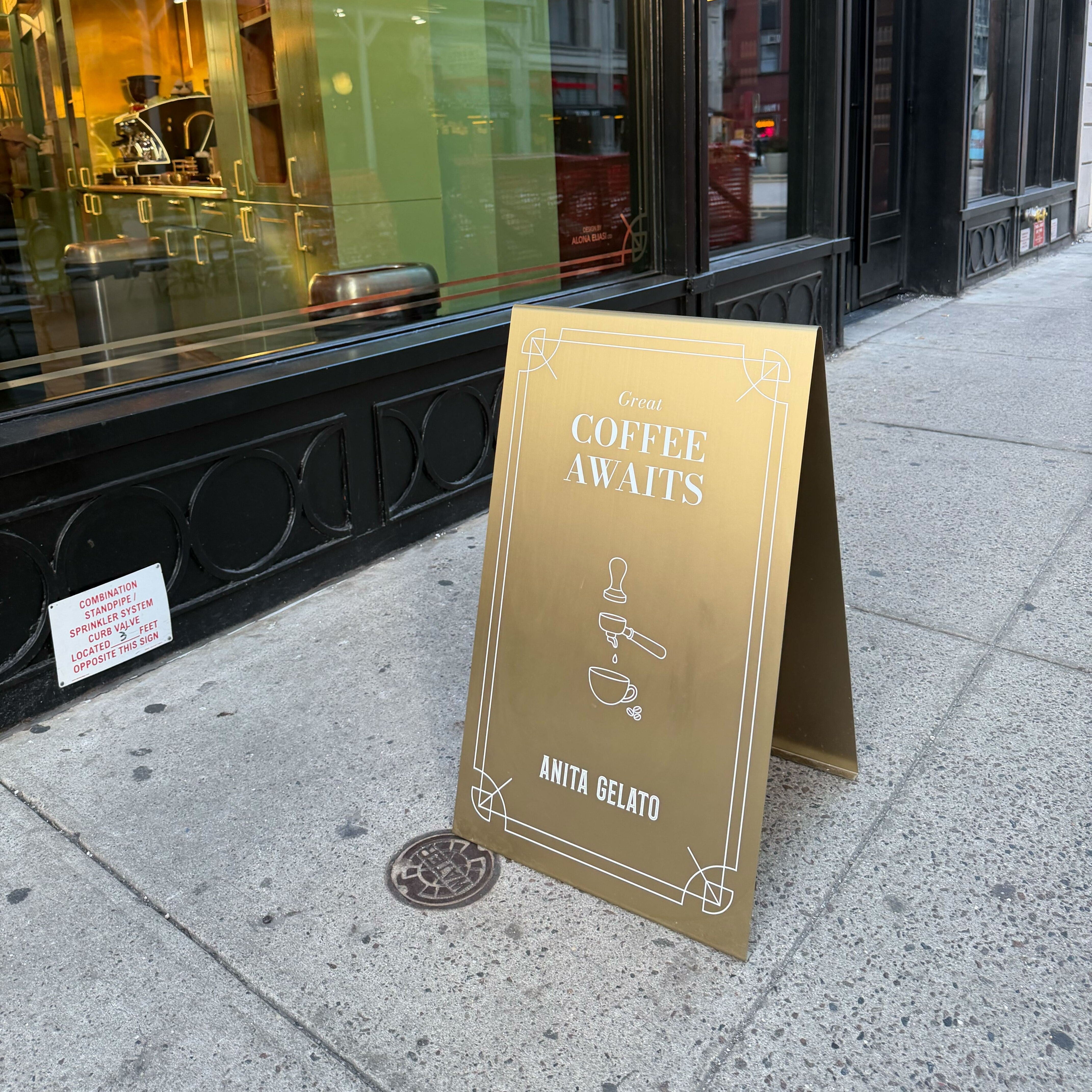Gold sign on a sidewalk in front of a store window with 'Coffee Awaits' text.