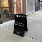 Black stainless steel A-frame sign with white Peachy lettering outside a storefront.