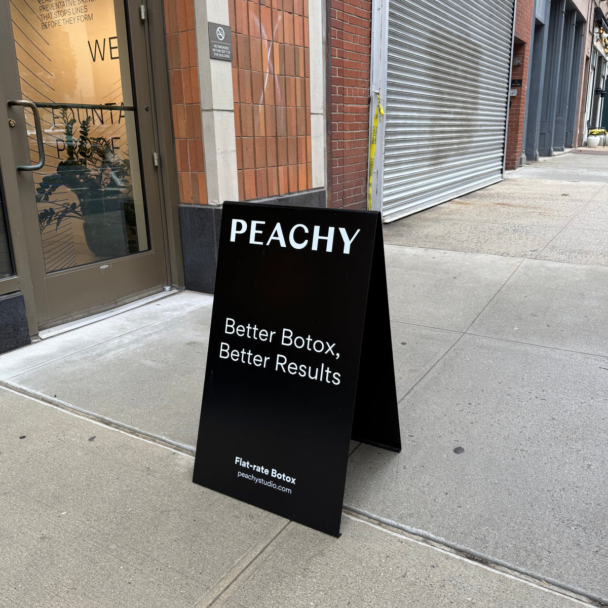 Black stainless steel A-frame sign with white Peachy lettering outside a storefront.